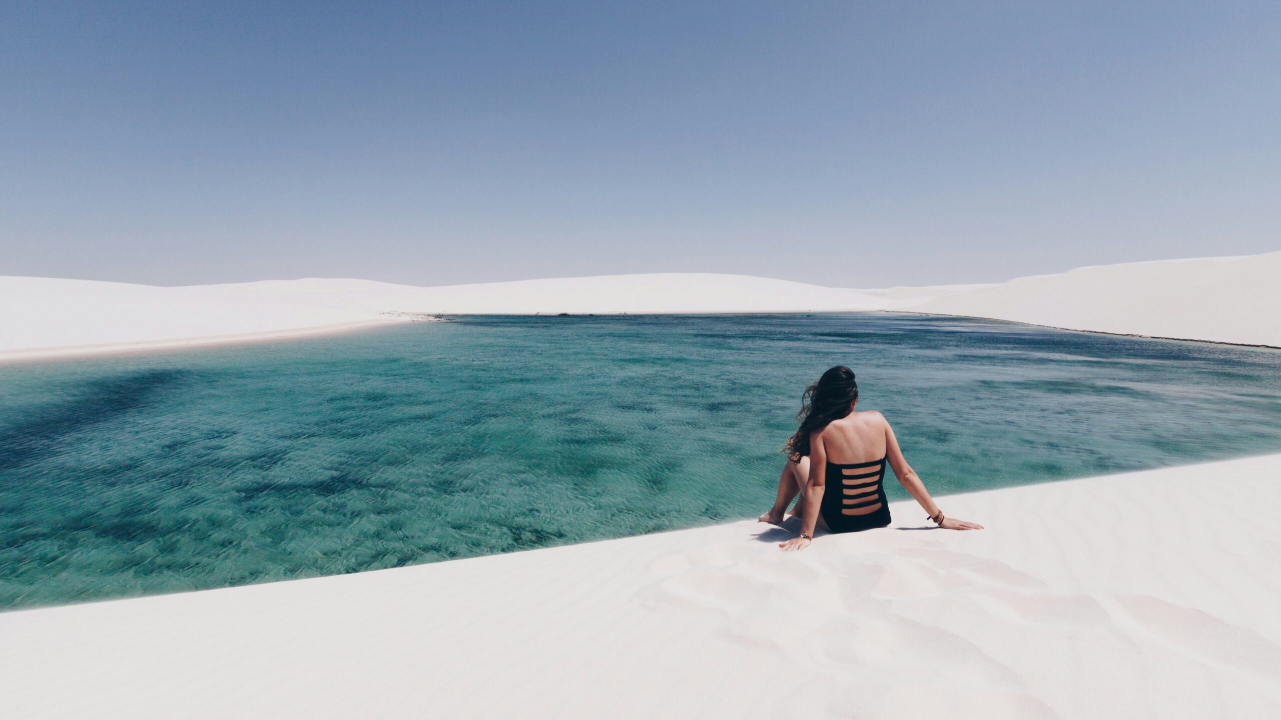 Woman sitting by serene turquoise water, dunes.